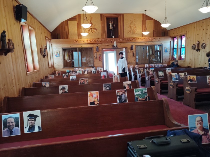 Small church with photos of parishioners taped to the pews when they couldn't come to Mass due to COVID. Altar boy near the doorway in white cassock. 